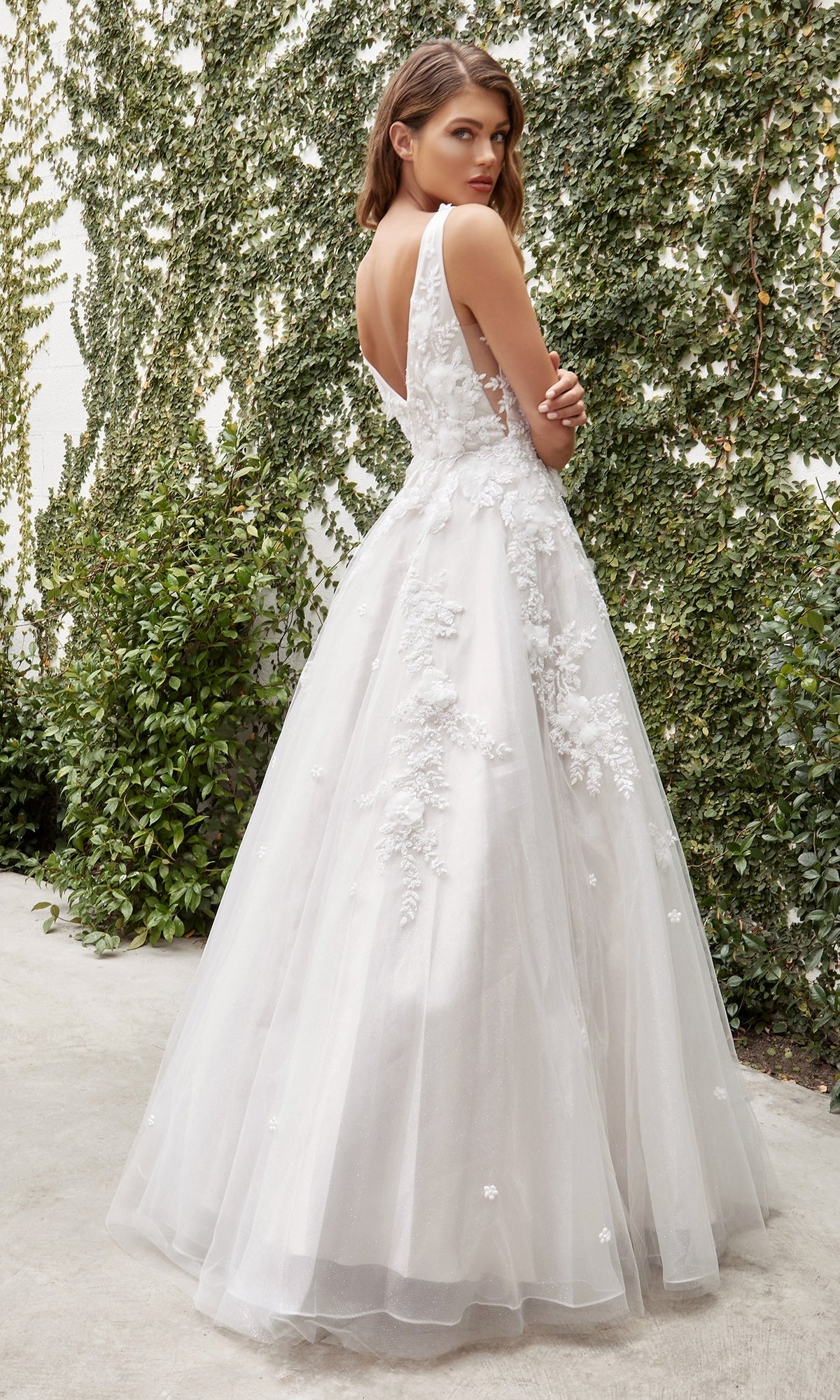A woman in the French White Long Wedding Gown, Ladivine A1028W, with lace floral details stands outdoors, looking over her shoulder amid lush green foliage.